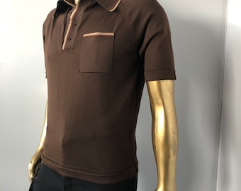 Könnte beinhalten: Ein braunes Poloshirt mit einer Brusttasche. Das Shirt hat einen Kragen und kurze Ärmel. Der Kragen und die Tasche sind mit einer helleren beigefarbenen Farbe verziert. Das Shirt wird mit einer schwarzen Hose getragen.