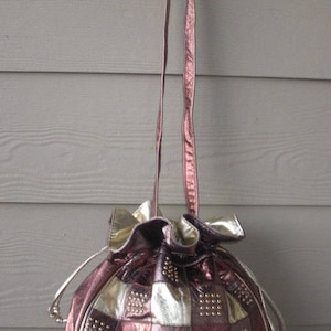 May include: A vintage drawstring handbag with a checkerboard pattern in metallic silver and burgundy. The bag features small gold studs and a long, thin strap. The bag is hanging against a gray wooden background.