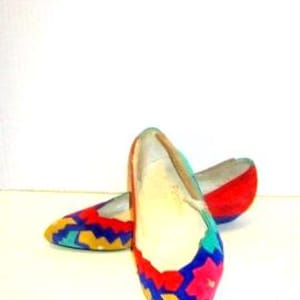 May include: A pair of colorful flats with a pointed toe. The shoes feature a vibrant pattern of blue, yellow, pink, and red geometric shapes. The shoes have a low heel and a soft, suede-like texture.
