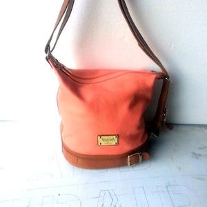 May include: Coral-colored shoulder bag with brown leather accents. The bag features a brown leather strap, base, and buckle. A gold-colored rectangular tag with the brand name is visible on the front of the bag. The bag is made of a soft, textured material.
