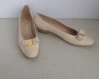 May include: A pair of beige women's flats with a bow detail and a small heel. The shoes have a textured appearance and a gold-colored buckle on the bow. The brand name "Salvatore Ferragamo" is visible inside one shoe.