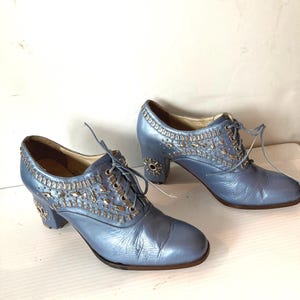 May include: A pair of light blue leather shoes with decorative silver embellishments. The shoes have a lace-up design and a small heel. The embellishments are concentrated around the laces and the heel.