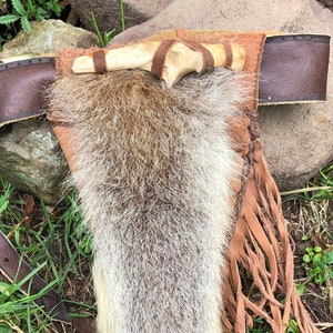 Può includere: Una cintura in pelle marrone con una borsa di pelliccia e un manico in osso. La borsa ha dettagli a frangia ed è decorata con un manico in osso.