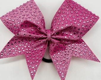 May include: A pink cheer bow with a sparkly finish and rhinestones. The bow is attached to a black elastic hair tie.