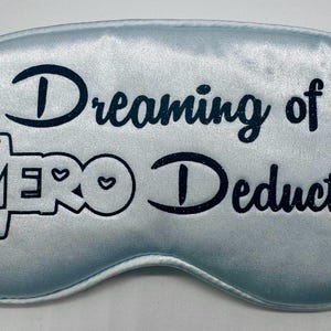 May include: A light blue sleep mask with the text "Dreaming of Zero Deductions" in black lettering. A small bow is in the top left corner.