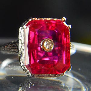 May include: A close-up of a vintage ring featuring a large, rectangular, vibrant red gemstone. The gemstone is set in a gold-toned bezel with a small, round, clear stone in the center. The band is silver-toned with intricate filigree detailing.
