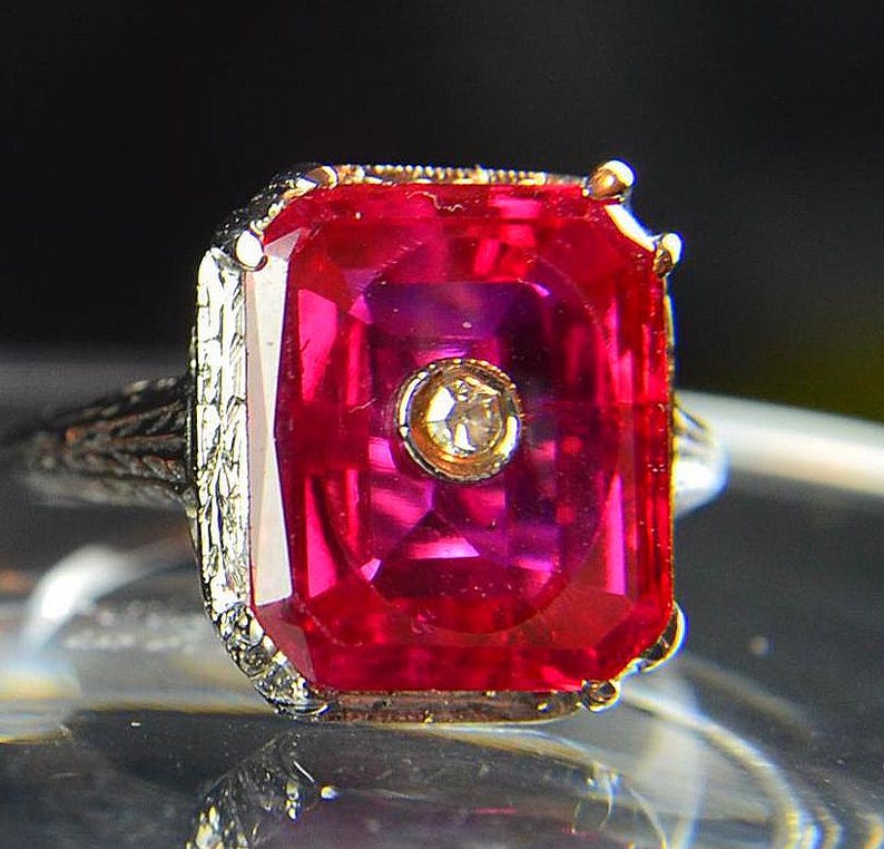 May include: A close-up of a vintage ring featuring a large, rectangular, vibrant red gemstone. The gemstone is set in a gold-toned bezel with a small, round, clear stone in the center. The band is silver-toned with intricate filigree detailing.