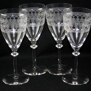 May include: A set of four clear glass wine glasses featuring a delicate etched lace-like pattern around the upper rim. Each glass has a slender stem with a small decorative knob and a wide circular base, standing upright on a dark surface.