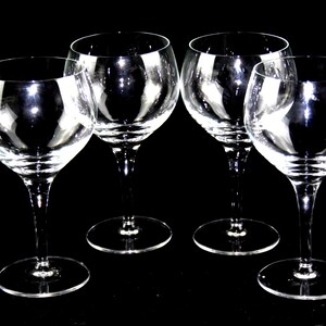 May include: A set of four clear glass wine glasses with rounded bowls, slender stems, and circular bases. Each glass features a smooth, transparent surface and a consistent design, arranged in a group against a solid black background.