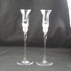 May include: A pair of clear glass candle holders with a simple, elegant design. The holders have a tall, slender stem and a flared base.