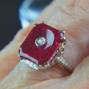 May include: A close-up of a vintage ring featuring a large, rectangular, deep red gemstone with a small, round, clear stone in the center. The ring has an ornate, filigree-style band.