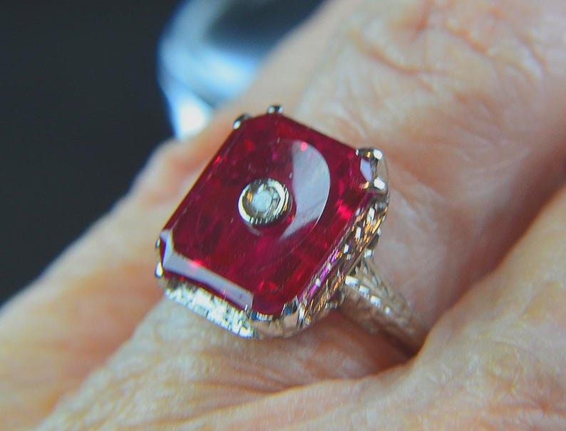 May include: A close-up of a vintage ring featuring a large, rectangular, deep red gemstone with a small, round, clear stone in the center. The ring has an ornate, filigree-style band.