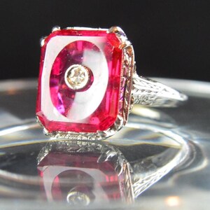 May include: A vintage ring featuring a large, rectangular, ruby-red gemstone with a white swirl design and a small diamond accent. The ring has an ornate, filigree-style silver band and setting. The ring is displayed on a reflective surface.