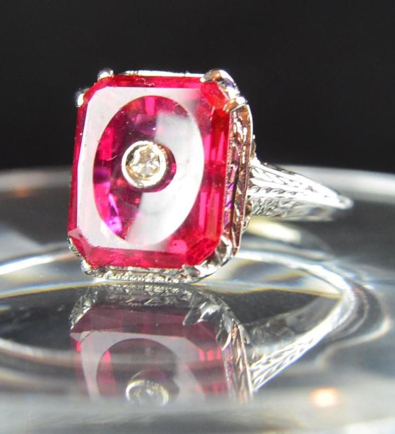 May include: A vintage ring featuring a large, rectangular, ruby-red gemstone with a white swirl design and a small diamond accent. The ring has an ornate, filigree-style silver band and setting. The ring is displayed on a reflective surface.
