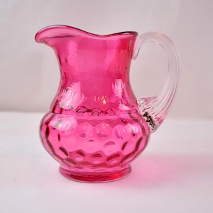 May include: A vibrant pink glass pitcher with a clear, curved handle. The pitcher has a bulbous body with a textured, bubble-like pattern. The spout is gently curved for pouring. A decorative and functional piece.