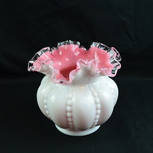 May include: A white milk glass vase with a pink ruffled top and silver trim. The vase has a round body with a beaded design.