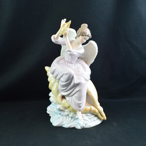 May include: A porcelain figurine of an angel sitting on a seashell. The angel is wearing a pink dress and has wings. The seashell is white with gold accents. The figurine is on a base that looks like waves.