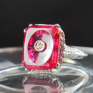 May include: A vintage ring with a rectangular, ruby-red gemstone and a small, round diamond at its center. The ring has an ornate, filigree-style band in a silver tone, showcasing intricate detailing. The gemstone is set in a silver-toned metal.