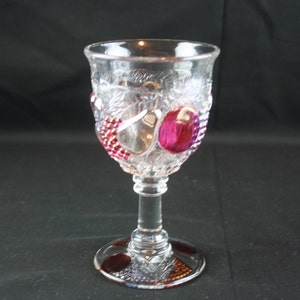 May include: A clear glass goblet with a stemmed base. The goblet is decorated with a raised design of grapes and leaves, and has pink and clear glass accents.