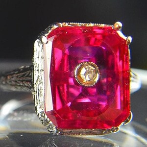 May include: A close-up of a vintage ring featuring a large, rectangular, ruby-red gemstone with a smaller, round, diamond-like stone in the center. The ring has an ornate, filigree-style band and gold accents.