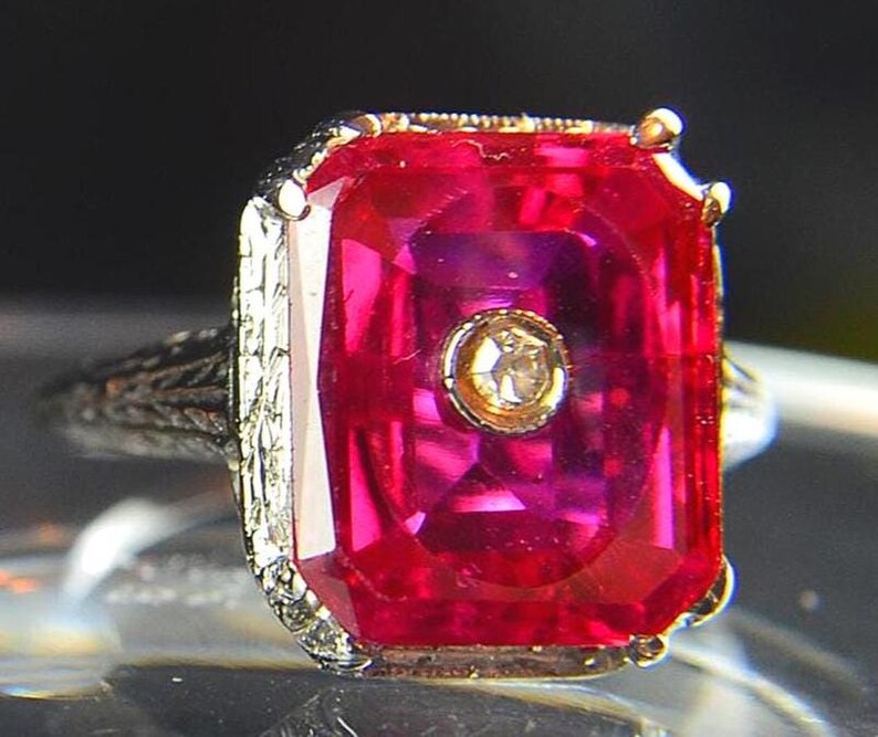 May include: A close-up of a vintage ring featuring a large, rectangular, ruby-red gemstone with a smaller, round, diamond-like stone in the center. The ring has an ornate, filigree-style band and gold accents.