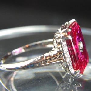 May include: A vintage-style ring with a large, rectangular, vibrant red gemstone. The ring features a detailed, ornate silver band with small accent stones around the main stone and on the band. The setting is intricate and elegant.