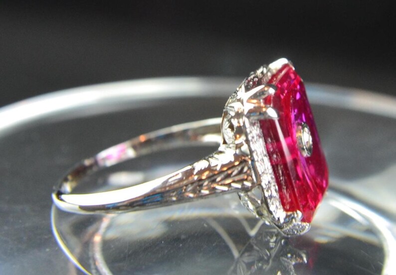 May include: A vintage-style ring with a large, rectangular, vibrant red gemstone. The ring features a detailed, ornate silver band with small accent stones around the main stone and on the band. The setting is intricate and elegant.