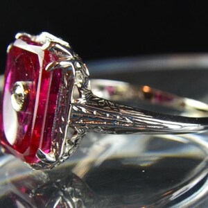 May include: A close-up of a silver ring featuring a large, rectangular, ruby-red gemstone with a small, round accent stone. The ring has intricate filigree detailing on the band and around the gemstone setting. The ring is displayed against a dark background.