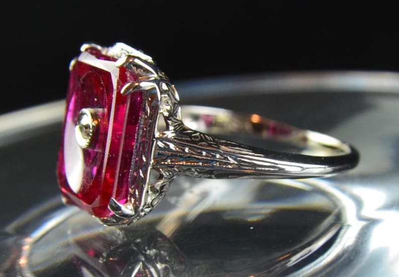 May include: A close-up of a silver ring featuring a large, rectangular, ruby-red gemstone with a small, round accent stone. The ring has intricate filigree detailing on the band and around the gemstone setting. The ring is displayed against a dark background.