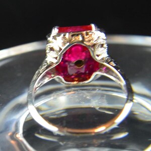 May include: A silver ring with a large, rectangular, red gemstone. The ring has intricate filigree detailing around the gemstone and on the band. The gemstone is set in a decorative setting, and the ring is displayed on a clear surface.
