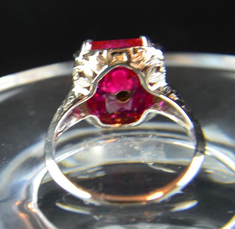 May include: A silver ring with a large, rectangular, red gemstone. The ring has intricate filigree detailing around the gemstone and on the band. The gemstone is set in a decorative setting, and the ring is displayed on a clear surface.