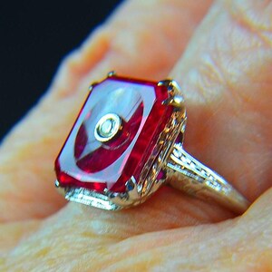May include: A close-up of a vintage ring featuring a large, rectangular, ruby-red gemstone with a central design. The ring has an ornate, filigree-style band in a silver tone, with small accent stones. The setting is detailed and elegant.