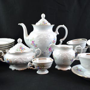 May include: A white porcelain tea set with floral accents. The set includes a teapot, sugar bowl, creamer, cups, saucers, and plates. Each piece has a gold trim and a delicate embossed design.