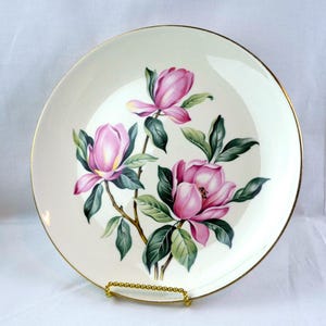 May include: A cream-coloured decorative plate with a floral design of pink magnolia blossoms and green leaves. The plate has a gold rim and is displayed on a gold-coloured stand. The floral design is centered on the plate.