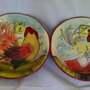 May include: Two ceramic rooster plates with colourful designs. The plates feature roosters with red, yellow, and blue feathers. The plates have a red rim and a green and yellow background with handwritten text.