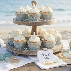 Può includere: Un'alzatina in legno a due livelli espone cupcakes con glassa bianca e decorazioni a forma di stella marina. I cupcakes sono in pirottini blu con motivo a pizzo. Un cartello recita "Dessert Table".