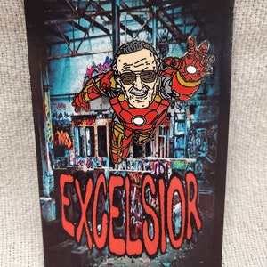 Puede incluir: Un pin de esmalte rojo, amarillo y dorado con una imagen de dibujos animados de Stan Lee como Iron Man. El pin está colocado sobre un fondo de una pared cubierta de grafitis con la palabra "EXCELSIOR" en letras rojas.