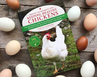 May include: A book titled "Keeping Healthy Chickens" with a white chicken on the cover. The book is surrounded by brown, white, and blue eggs on a wooden surface.