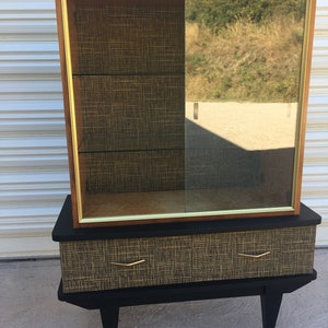 Peut inclure: Un meuble de style moderne du milieu du siècle avec une porte en verre et deux tiroirs. Le meuble est noir avec des garnitures dorées et présente un motif tissé sur le devant.