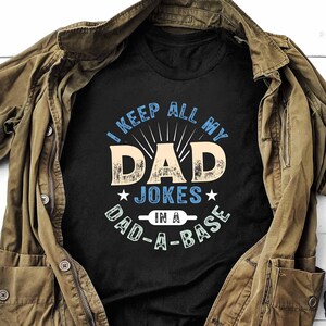 May include: A black t-shirt with a blue and white graphic that says "I keep all my dad jokes in a dad-a-base". The shirt is laid out on a white wooden surface with a brown jacket and brown boots.