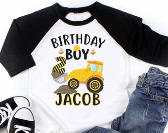 May include: A white and black raglan-style shirt with the text "BIRTHDAY BOY JACOB" and a yellow construction vehicle graphic. The shirt is paired with blue jeans and black sneakers.