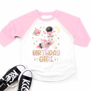 May include: White and pink raglan-style t-shirt with a space-themed graphic. The design includes an astronaut, rocket, planets, and the text "BIRTHDAY GIRL". Black and white sneakers are at the bottom of the image.