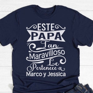 May include: Navy blue t-shirt with white text. The text reads "ESTE PAPA Tan Maravilloso Le Pertenece a Marco y Jessica" in a decorative font. The shirt is folded with a pair of blue jeans.