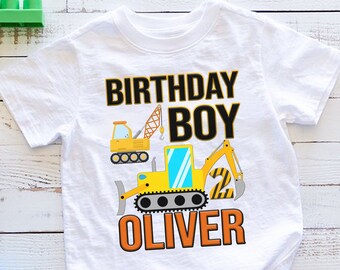 May include: White short-sleeved t-shirt featuring a colorful construction vehicle design. The shirt has the words "BIRTHDAY BOY" in black and orange, the name "OLIVER" in orange, and the number "2". The shirt is made of a soft material.