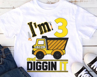 May include: White t-shirt with a construction-themed design. The shirt features the text "I'm 3" with a hammer and saw graphic, a yellow dump truck, and the words "Jacob" and "and Diggin It". Blue jeans and black and white sneakers are also in the image.