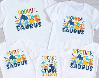 Może przedstawiać: Pięć białych koszulek z grafiką dinozaurów i tekstem. Koszulki mówią "Daddy...saurus", "Mommy...saurus", "Sister...saurus", "Brother...saurus" i "Roar I'm a Twol-A-saurus".