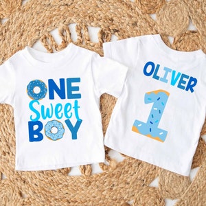 May include: Two white t-shirts with blue and white donut designs. One shirt says "One Sweet Boy" and the other says "Oliver" with the number "1".