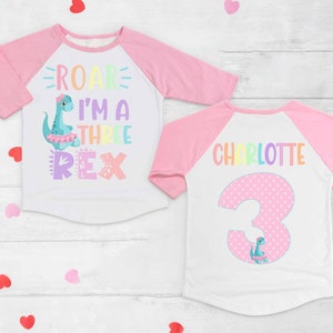 May include: A pink and white long-sleeved shirt with a rainbow dinosaur graphic and the text "Roar I'm a Three Rex". The back of the shirt has a pink polka dot number 3 with a dinosaur graphic and the name "Charlotte".