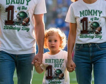 May include: A family of three wearing matching white t-shirts with football-themed graphics. The shirts read "DADDY of the 1st Birthday Boy," "MOMMY of the 1st Birthday Boy," and "BIRTHDAY BOY 1st Jacob." The family is smiling.
