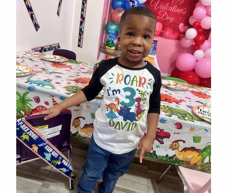 May include: A young boy wearing a white and black baseball shirt with the text "Roar I'm 3 David" in colorful letters. He is standing in front of a table with a dinosaur-themed tablecloth.